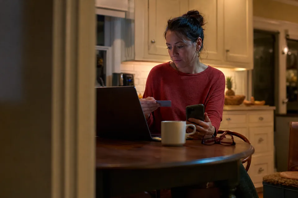 Woman at a kitchen table with a credit card and phone, reviewing a monthly membership charge on her laptop